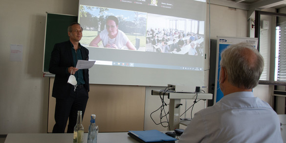 On the left in the photo is Prof. Cornelius Schubert. Prof. Johannes Weyer is sitting in front on the right with his back to the camera. A screen can be seen in the background. On the right side of the screen, you can see Prof. Cornelia Kropp, who is digitally connected. On the right side of the screen, you can see the camera shot of the auditorium.