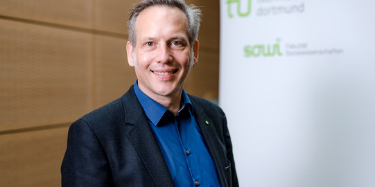 Prof. Dr. Cornelius Schubert The photo shows the portrait of Prof. Cornelius Schubert. Behind him is a rollup on which the logos of the TU Dortmund University and the Department of Social Sciences can be read.