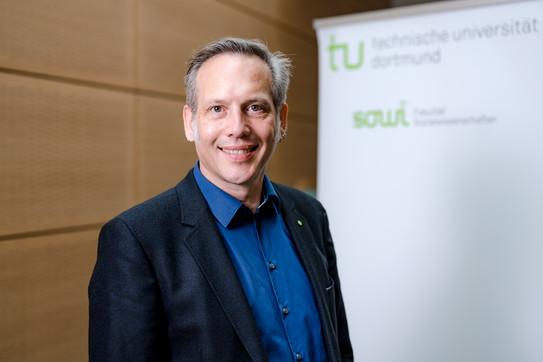 The photo shows the portrait of Prof. Cornelius Schubert. Behind him is a rollup on which the logos of the TU Dortmund University and the Department of Social Sciences can be read.