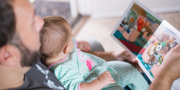 Ein Vater hat ein Baby auf dem Schoß und schaut mit ihm ein Bilderbuch an.