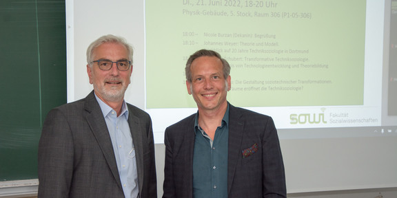 Auf dem Foto steht rechts Prof. Dr. Johannes Weyer. Links neben ihm steht Prof. Dr. Cornelius Schubert. Im Hintergrund ist auf einer Leinwand das Programm der gemeinsamen Abschieds- und Willkommensveranstaltung zu sehen.
