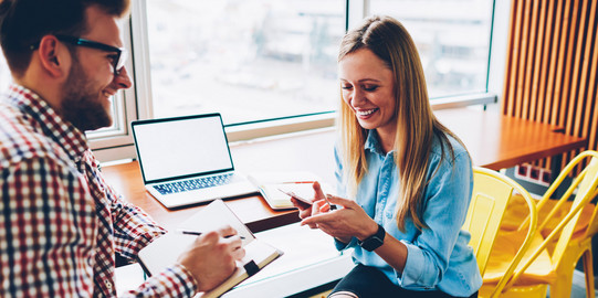 a man and a woman are sitting at a desk facing each other. On the desk is an open laptop
