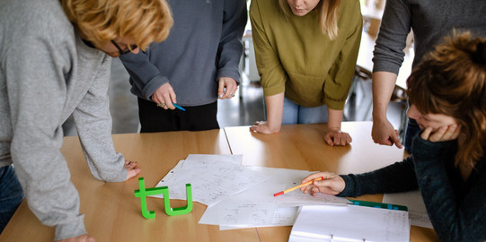 Studierende Gruppenarbeit fünf Studierende stehen um einen Tisch und lesen ein Dokument.