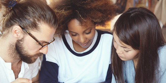 Studierende mit Unterlagen Drei Studierende haben ihre Köpfe über Unterlagen zusammengesteckt.