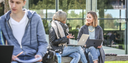 Zwei Studentinnen unterhalten sich draußen. Sie haben Laptops auf dem Schoß.