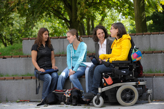 Studierende mit Behinderung Vier Studierende, darunter Studierende mit Behinderung, sitzen gemeinsam auf dem Campus.
