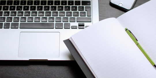 Laptop und Notizbuch mit Stift auf einem Tisch liegend.