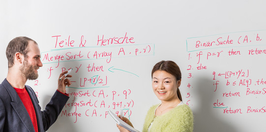 Two people in front of a whiteboard. One is holding a red pen, the other a notepad.