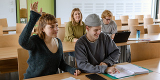 Studierende im Seminarraum sitzen an den Tischen. Eine Person meldet sich. 