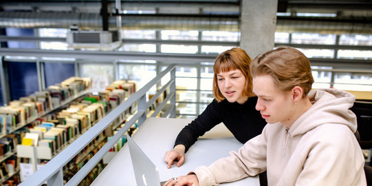 Arbeiten in der Bibliothek Zwei Studierende sitzen lernend vor einem Laptop in der Bibliothek.
