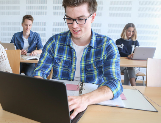 Student sitzt im Seminarraum am Laptop, im Hintergrund weitere Studierende