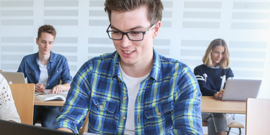 Studierender mit Laptop Student sitzt im Seminarraum am Laptop, im Hintergrund weitere Studierende