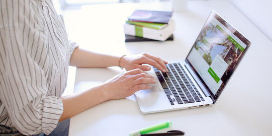 Studentin tippend am Laptop, daneben liegen Bücher und Stifte.