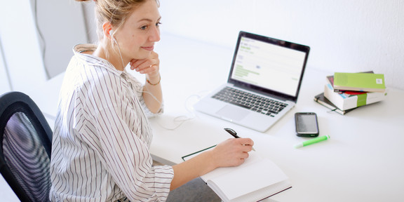 Frau sitzt mit Laptop am Schreibtisch und notiert etwas in ein Notizbuch