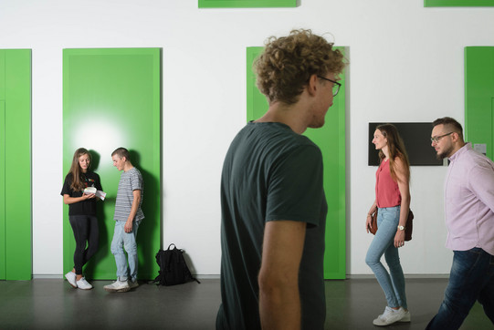 Studierende im Gehen Zwei Studierende lehnen an der Wand, zwei kommen von rechts ins Bild und einer geht nach rechts.